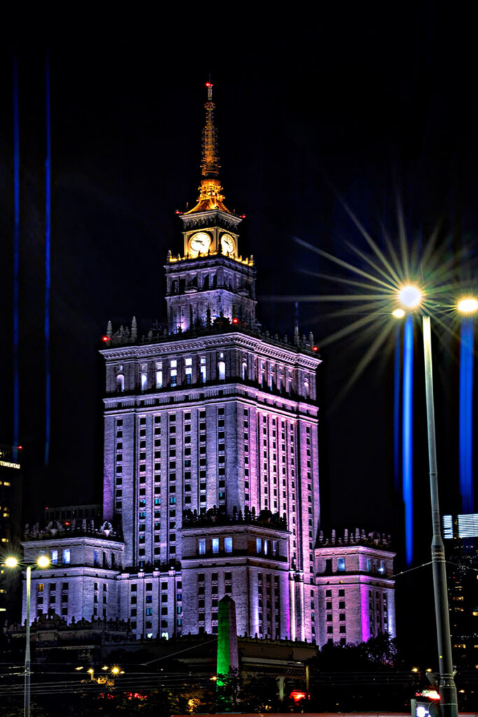 Das Bild zeigt den Kulturpalast in Warschau bei Nacht, eindrucksvoll in violettem Licht angestrahlt. Durch den Einsatz eines anamorphischen Objektivs entstehen markante blaue Lichtstreifen, die von der hellen Straßenbeleuchtung erzeugt werden und der Szene eine filmische, urbane Tiefe verleihen.