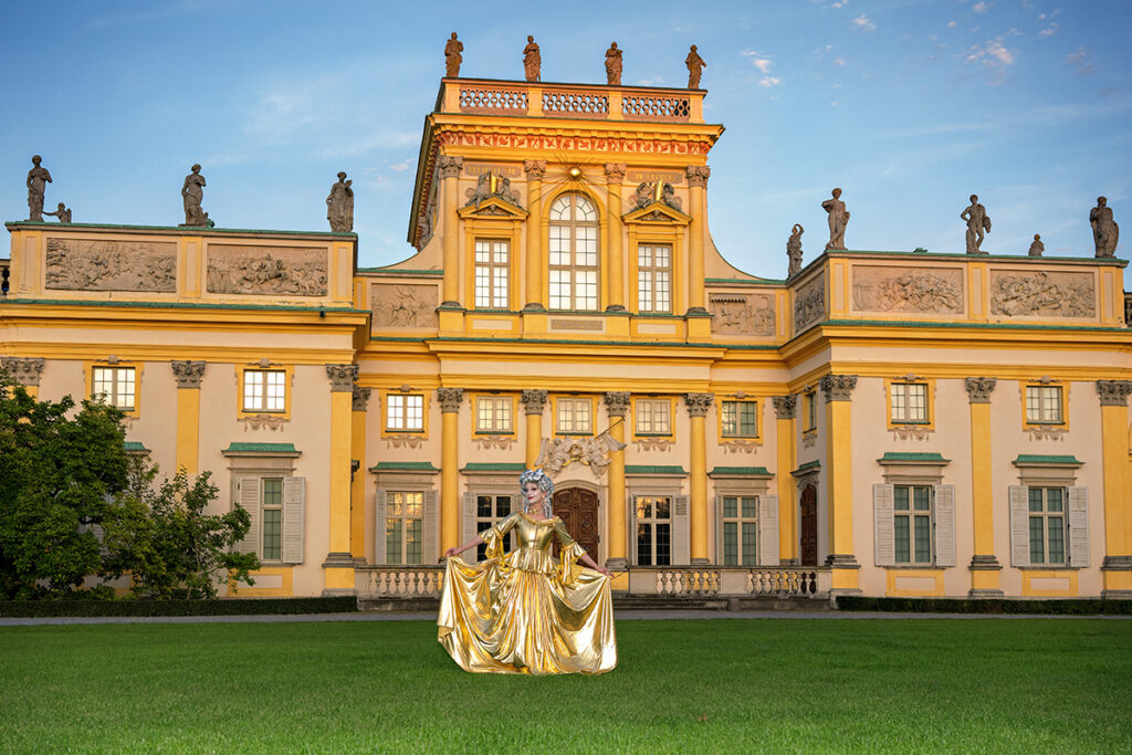 Auf dem Bild ist unser Model Nathalie zu sehen, posierend auf dem Rasen vor dem historischen Schloss Wilanów in Warschau (Polen). Die Dämmerung hat bereits eingesetzt und taucht die Szene in ein weiches, atmosphärisches Licht. Nathalie trägt ein goldenes Kleid und eine barocke Perücke im Stil von Marie Antoinette, wodurch eine eindrucksvolle Verbindung aus historischer Inszenierung und moderner Fotografie entsteht.